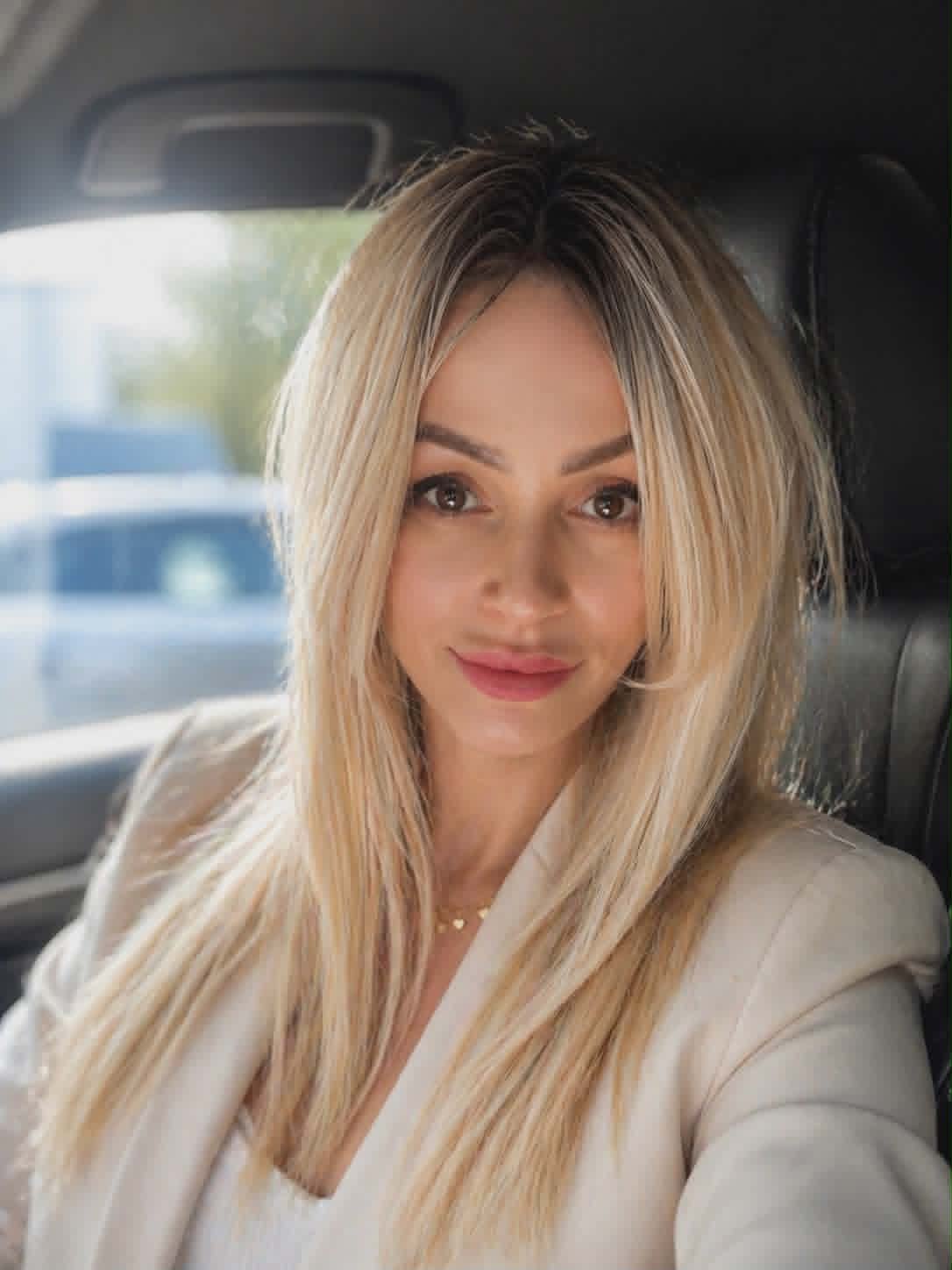 Woman with long blonde hair and pink lipstick sitting in a car, looking at camera